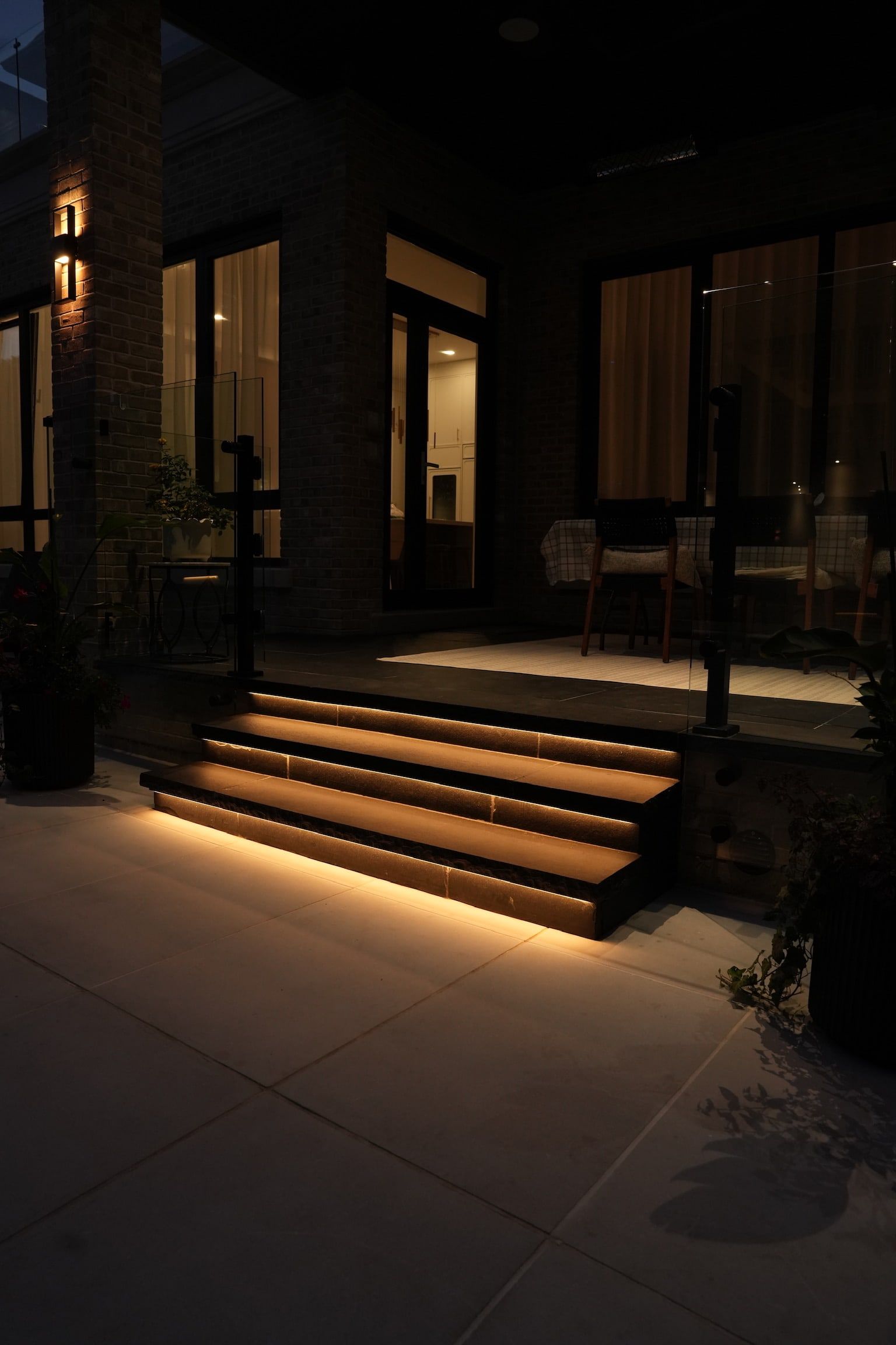 Porch steps at night with warm light under each step, beside a dark brick wall.