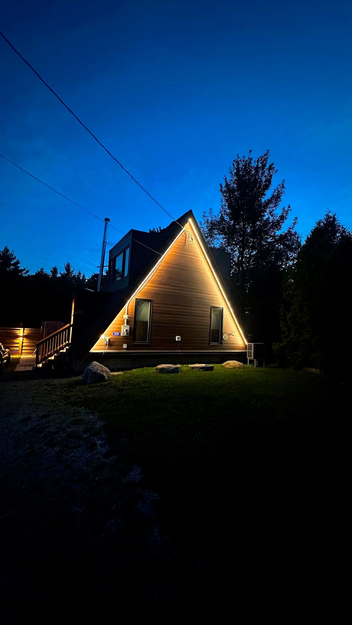 An A-frame cottage at dusk with warm light tracing its roof edges, among tall trees.
