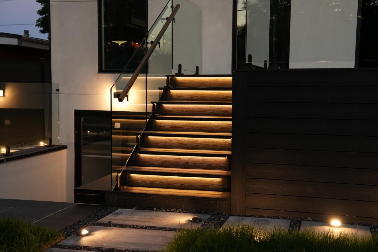 An exterior stair with a glass railing and dark wood cladding at dusk, with warm light under each tread.