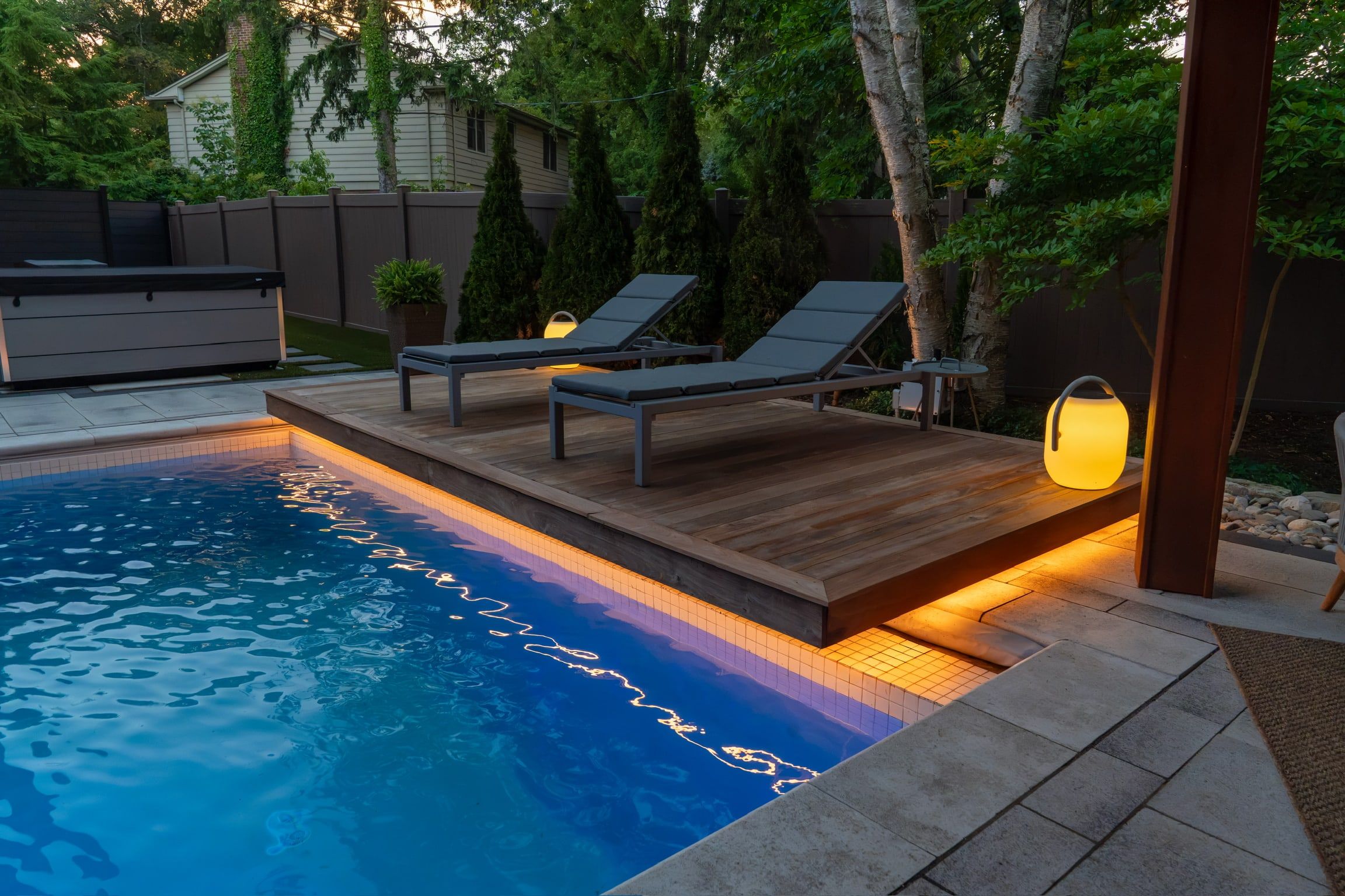 A raised timber deck beside a pool at dusk with warm light under its edge, loungers on top.