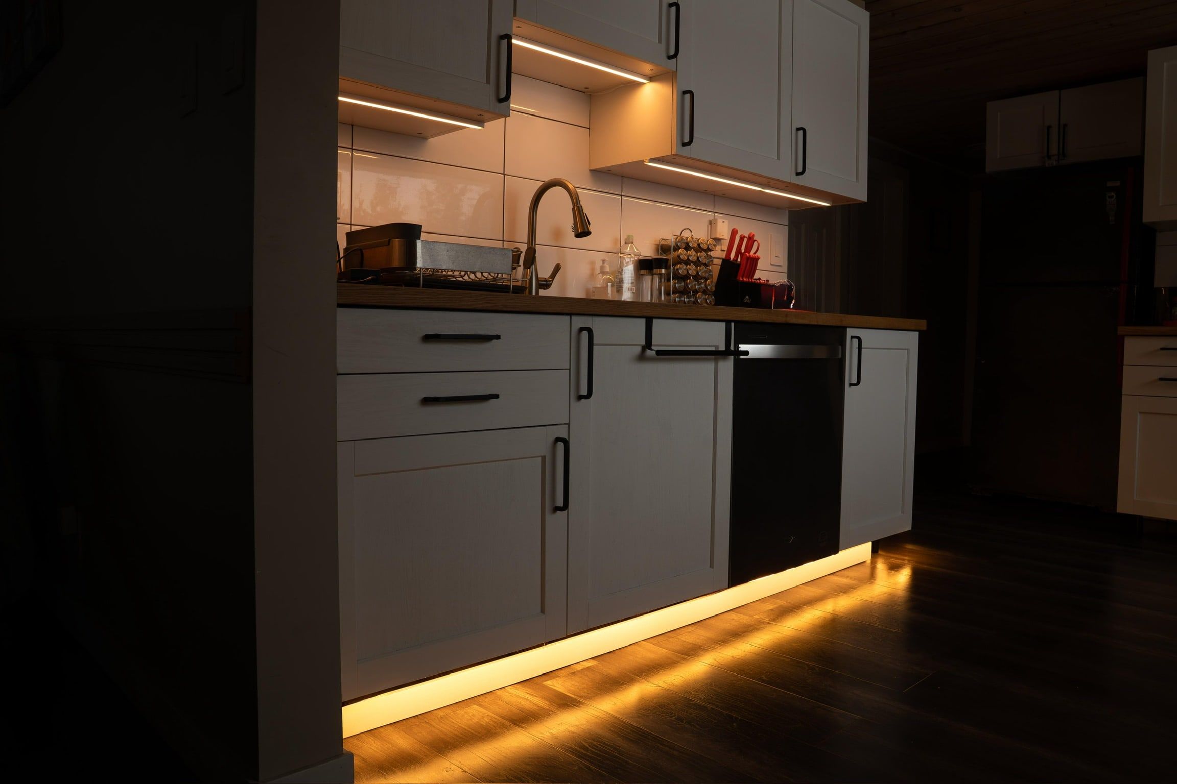 A white kitchen at night with warm light under the upper cabinets and a light line along the toe-kick.