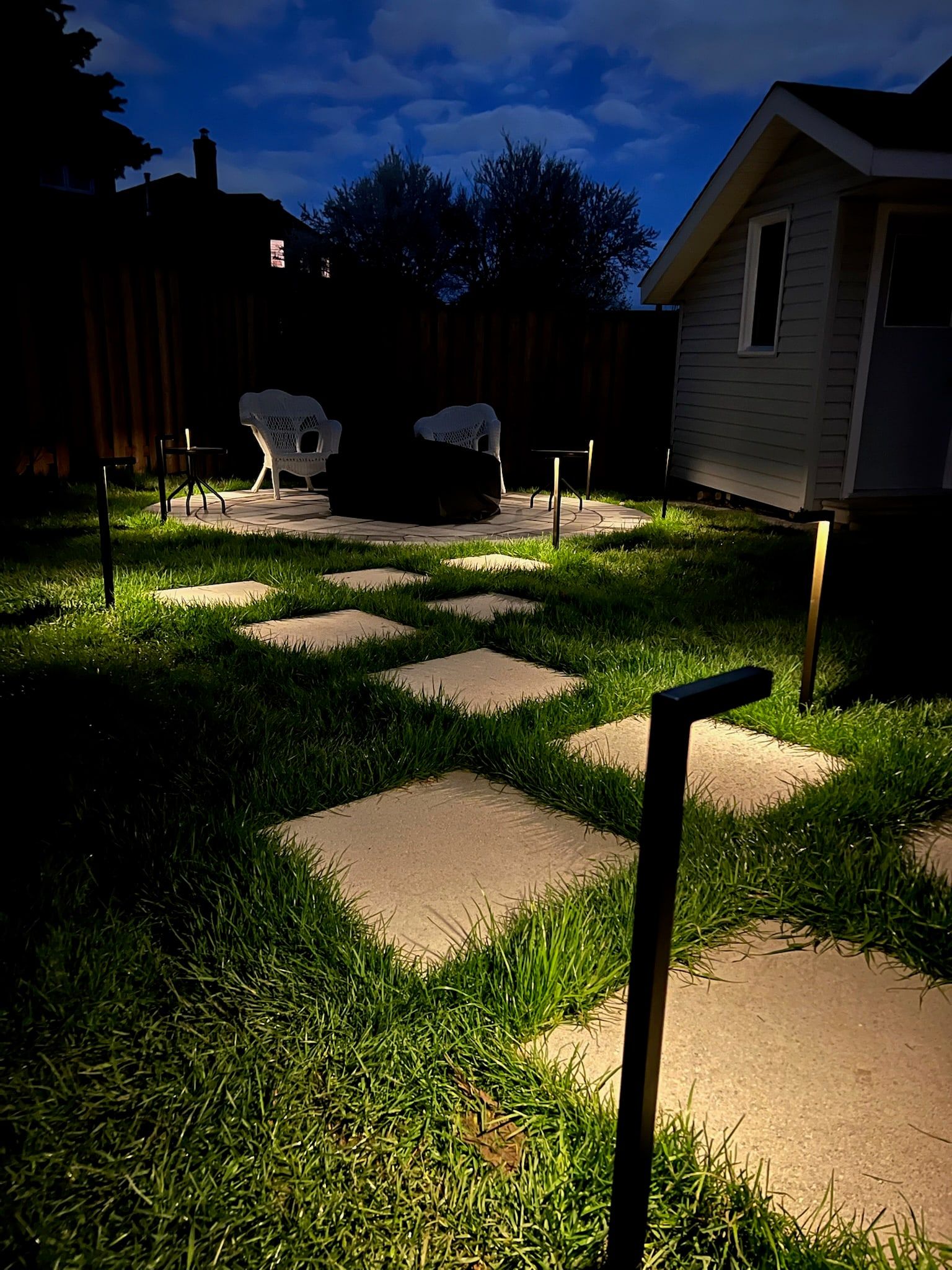 Square stepping stones across a lawn at dusk, lit by low path lights, leading to a round paver patio.