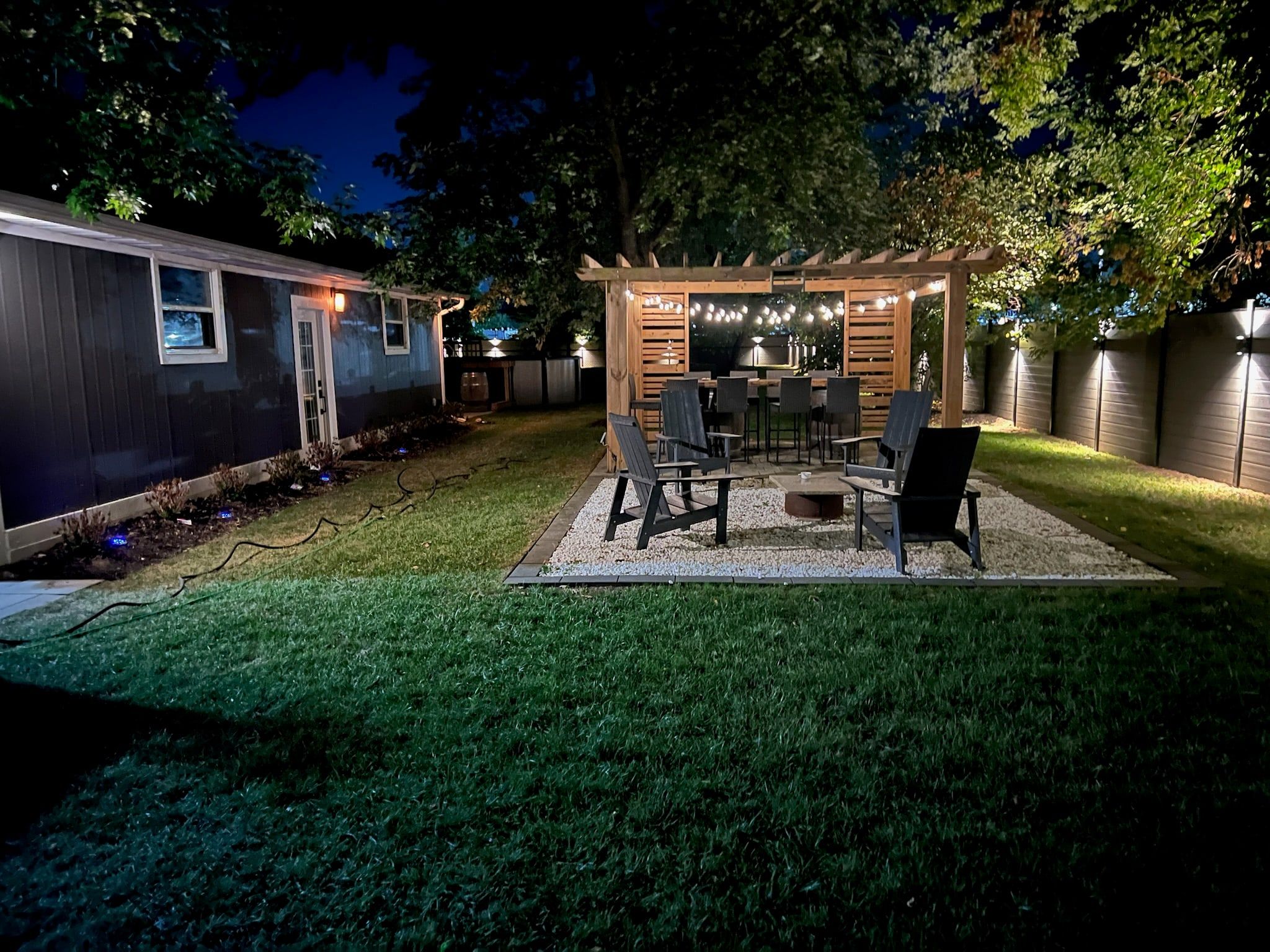 A backyard at night with a cedar pergola over a gravel patio, framed by a lit grey fence and uplit trees.