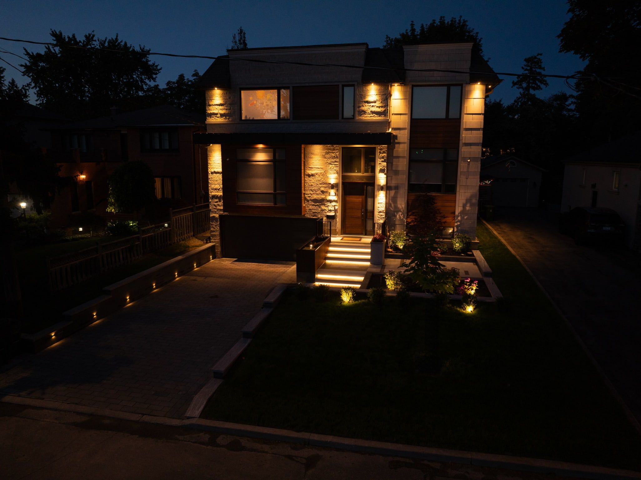 Aerial view at dusk of a modern two-storey home with lit front steps, path lights and an uplit tree.