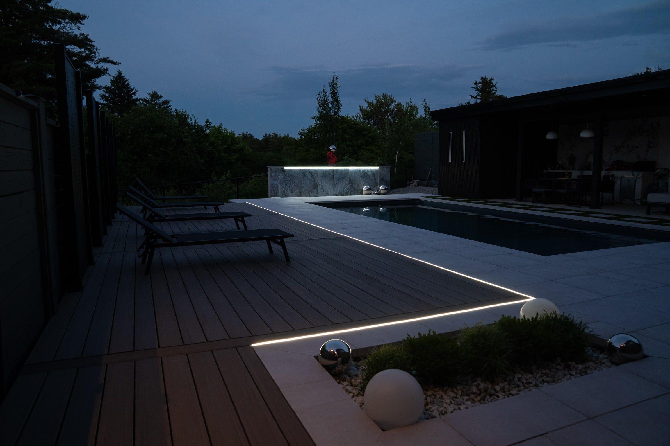 A dark composite pool deck at dusk with a continuous light line along its edge and lounge chairs beside the pool.