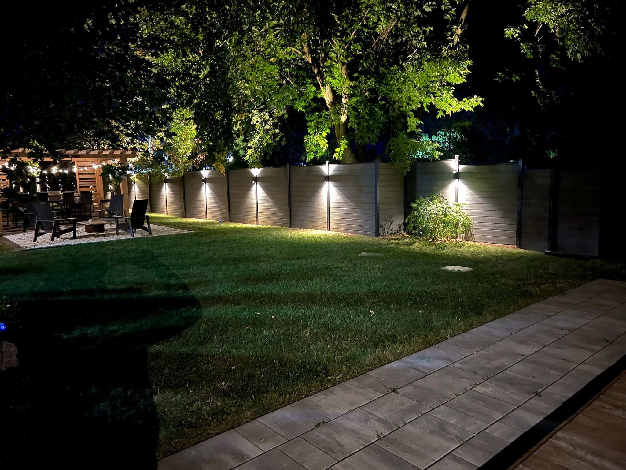 The lawn and paver path at night with the fence line lit.