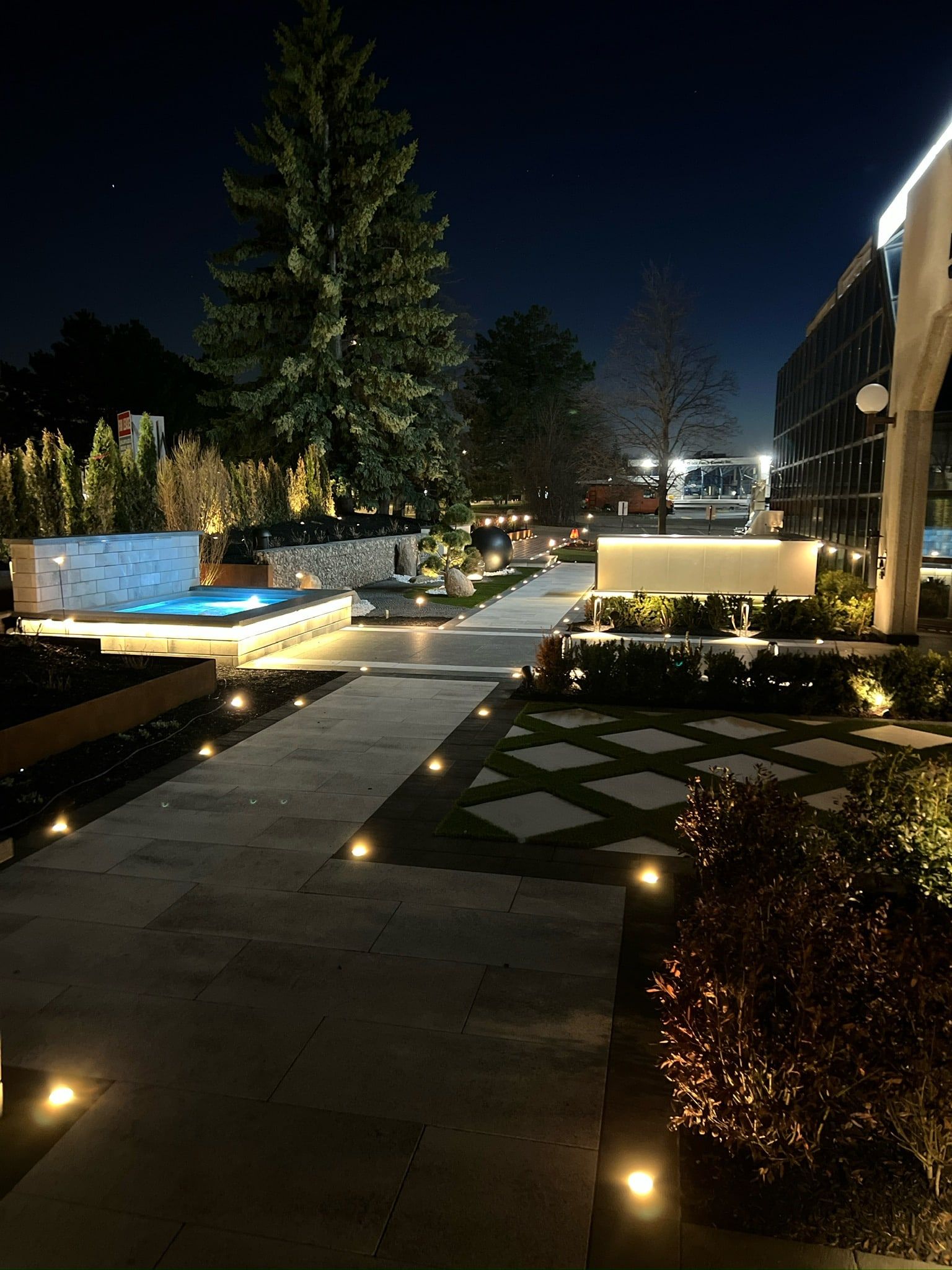 Square pavers set in the lawn, lit at night, with an evergreen behind.
