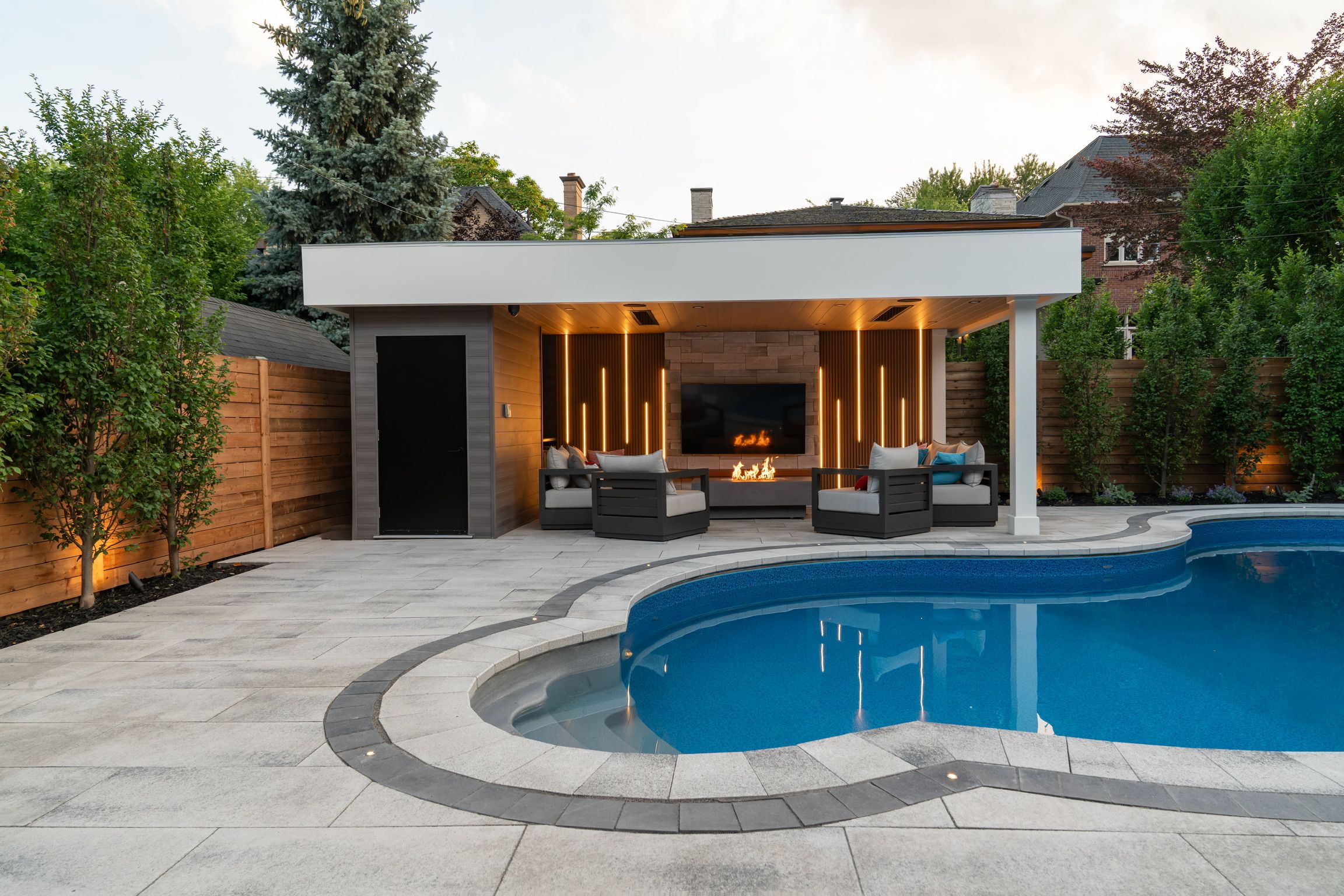 A white-roofed pool cabana at dusk with vertical warm light lines in wood slat walls either side of a stone fireplace, reflected in the pool.